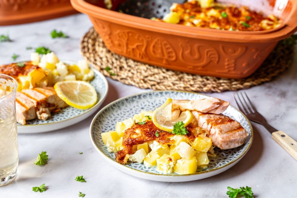 Bild 1: Kohlrabi-Gratin mit gegrilltem Lachs und Zitronenscheibe auf gemustertem Teller, im Hintergrund Terrakotta-Römertopf mit restlichem Gratin