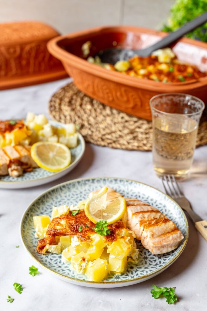 Bild 2: Angerichteter Teller mit Kohlrabi-Gratin und Lachs neben Weinglas, im Hintergrund geöffneter Römertopf und zweiter Teller