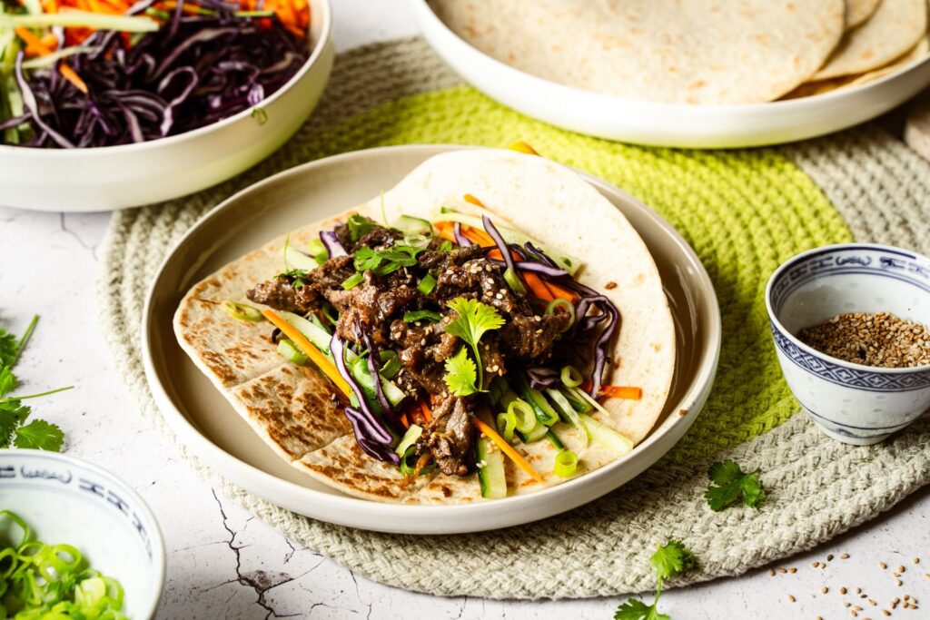Bild 1: Korean BBQ Wrap mit Sticky Bulgogi, Rotkohl, Möhren und Gurke auf weißem Teller, garniert mit Koriander und Sesam