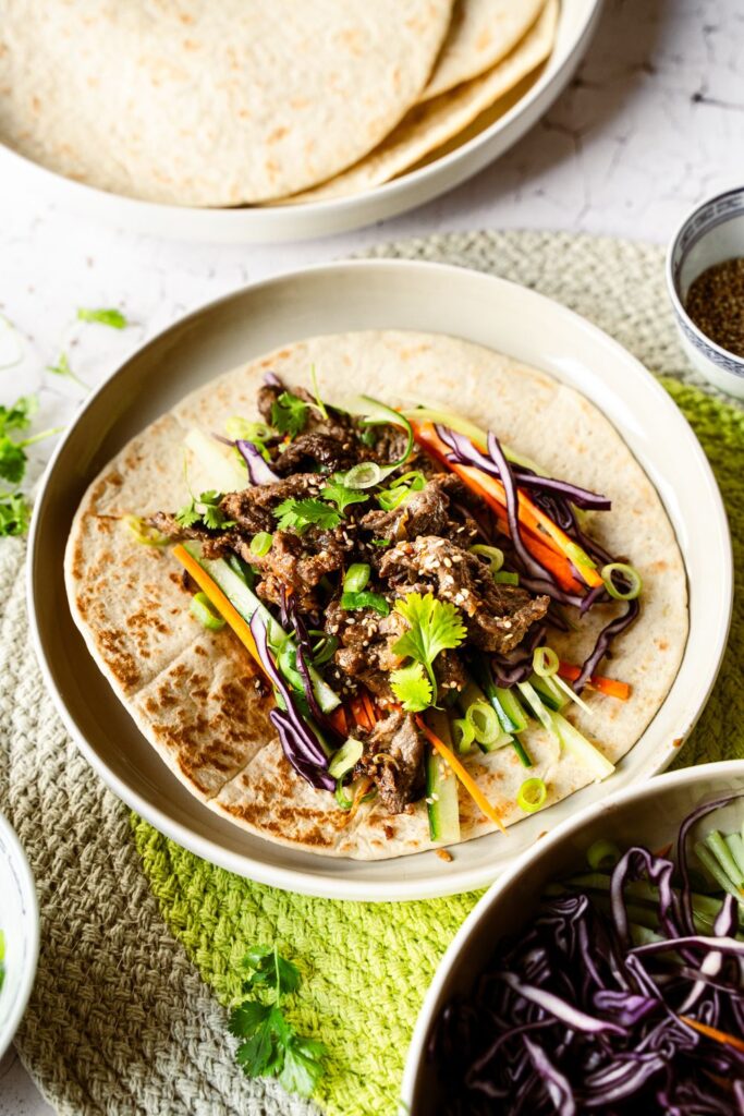 Bild 2: Angerichteter Bulgogi Wrap mit buntem Gemüse auf Teller, im Hintergrund Wraps und Schale mit Rotkohl-Streifen
