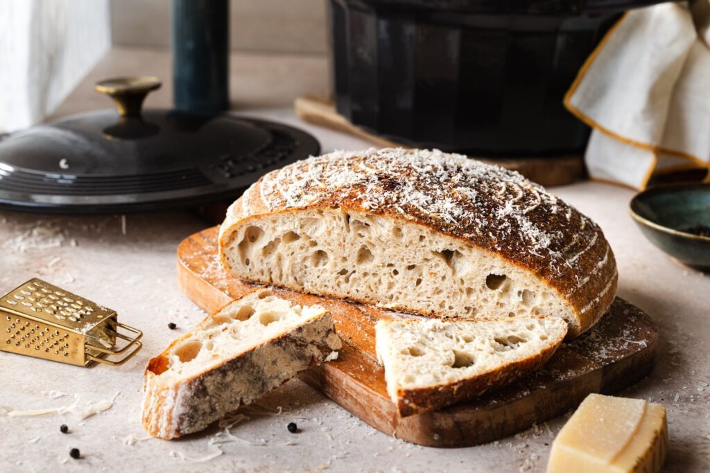 Aufgeschnittenes Pfeffer-Parmesan Brot mit Parmesankruste auf Holzbrett, im Hintergrund geöffneter Gusseisentopf mit Deckel