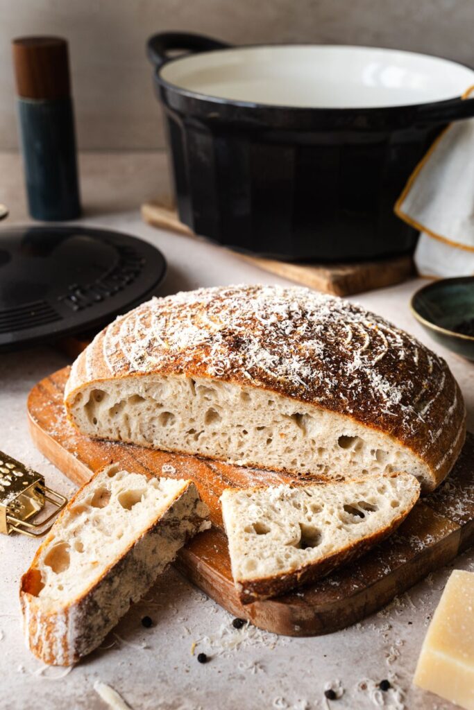 Angeschnittenes Sauerteigbrot mit Parmesan und Pfeffer auf Holzbrett, daneben Parmesanstück und Käsereibe