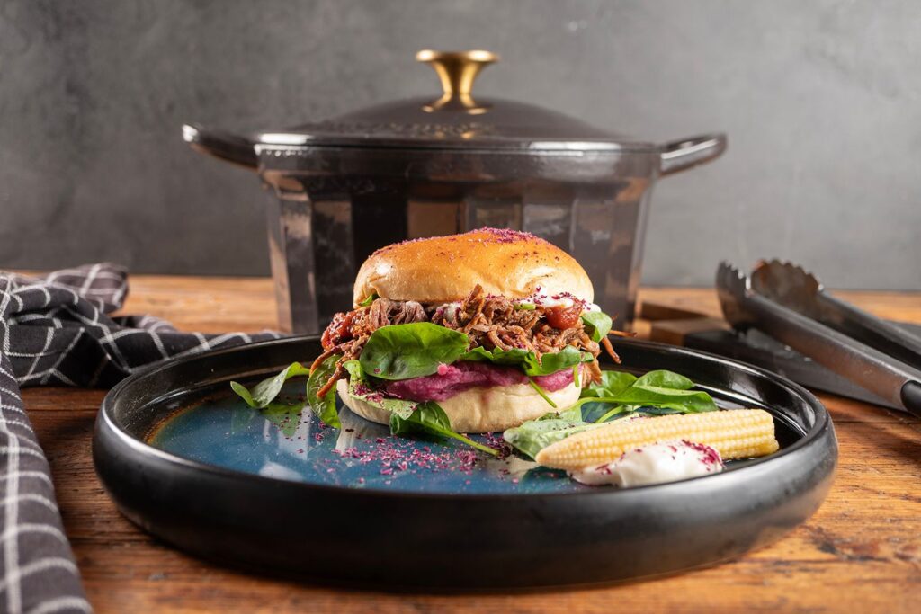 Pulled Beef Burger mit lila Süßkartoffelpüree und Spinat auf dunklem Teller von Moritz und Moritz, im Hintergrund gusseiserner Topf