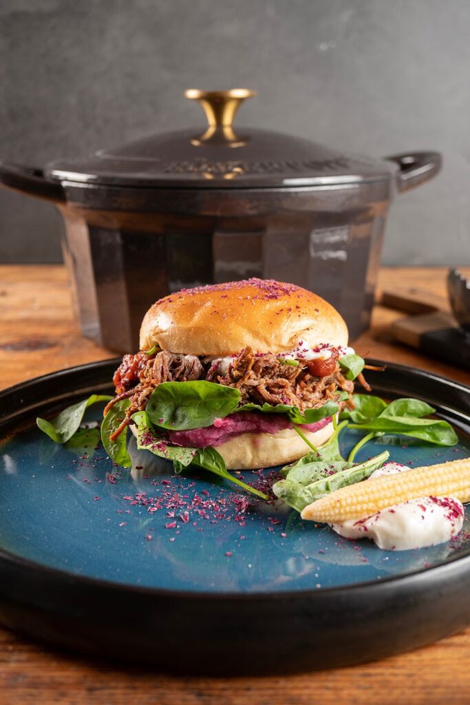 Angerichteter Pulled Beef Burger mit Baby-Maiskolben und Aioli auf blauer Schale, im Hintergrund geschlossener Gusseisentopf