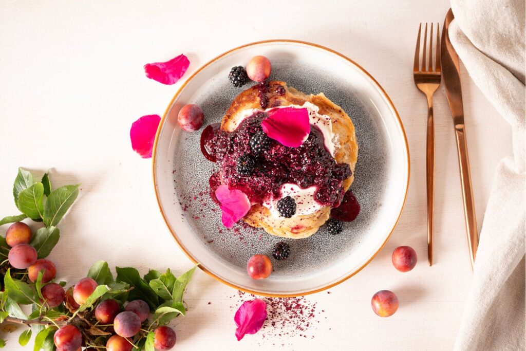 Sauerteig Pancakes mit Schmand, warmen Brombeeren und Rosenblättern auf grau-blauem Teller, umgeben von frischen Pflaumen