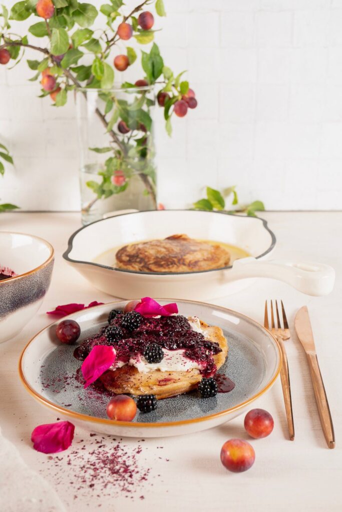 Sauerteig Pancake in weißer Gusseisenpfanne im Hintergrund, davor angerichteter Teller mit Brombeeren und Schmand