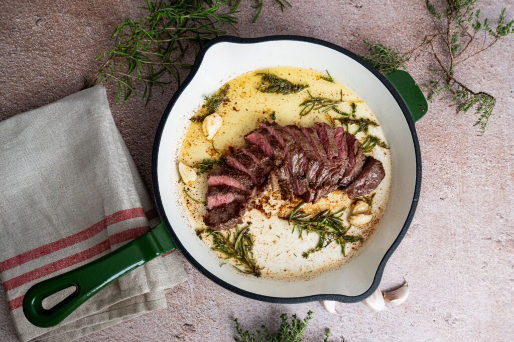 Aufgeschnittenes Steak in grüner Gusseisenpfanne mit Rosmarin, Thymian und Knoblauchzehen in geschmolzener Butter