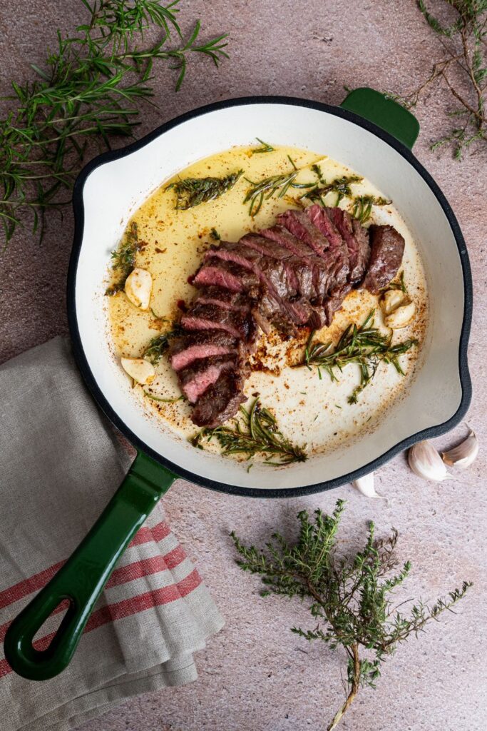 Angeschnittenes Steak in Gusseisenpfanne von oben, umgeben von frischen Kräuterzweigen und Knoblauch