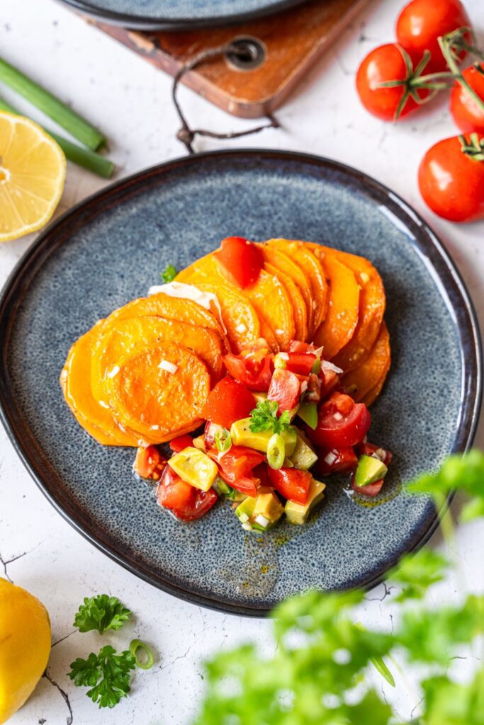 Angerichteter Teller mit Süßkartoffelscheiben und frischer Avocado-Tomaten-Salsa, im Hintergrund Zitrone und Kirschtomaten