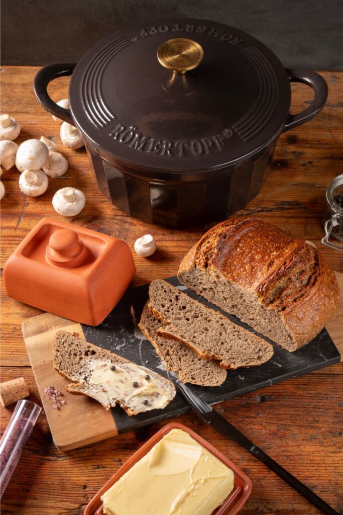 Champignon-Sauerteigbrot und Butterdose vor schwarzem Gusseisentopf, umgeben von frischen Champignons und Pfefferkörnern