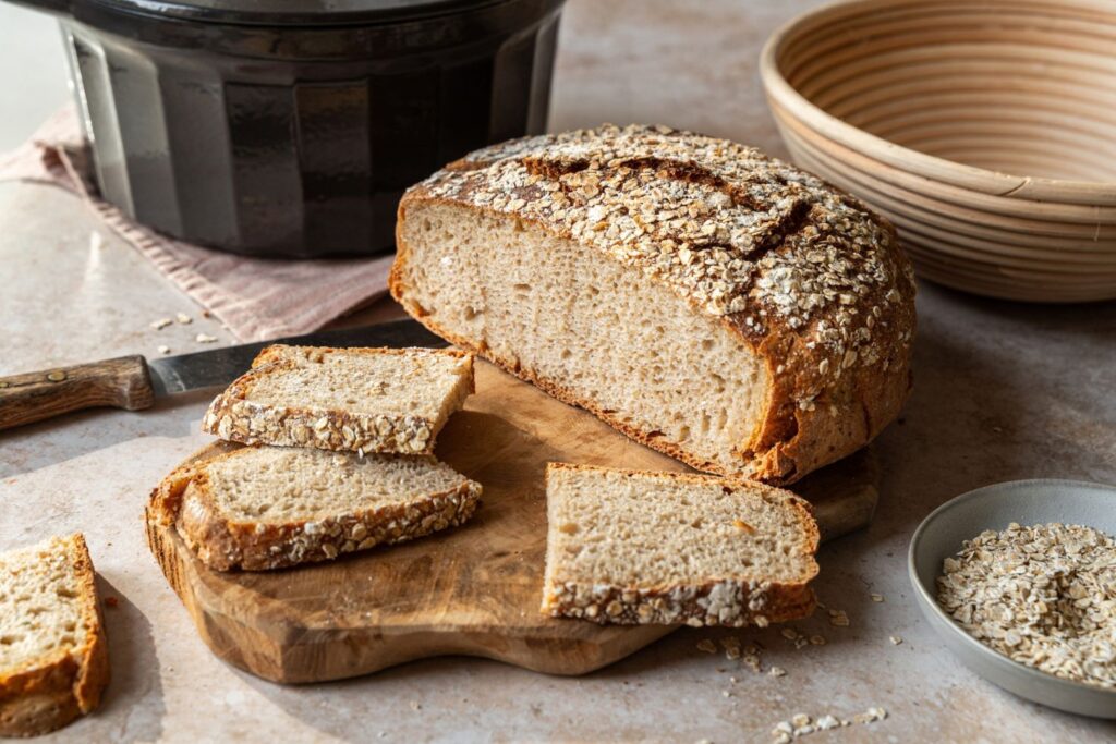 Aufgeschnittenes Hafer-Porridge Brot mit Haferkruste auf Holzbrett, im Hintergrund schwarzer Gusseisentopf und Gärkörbchen