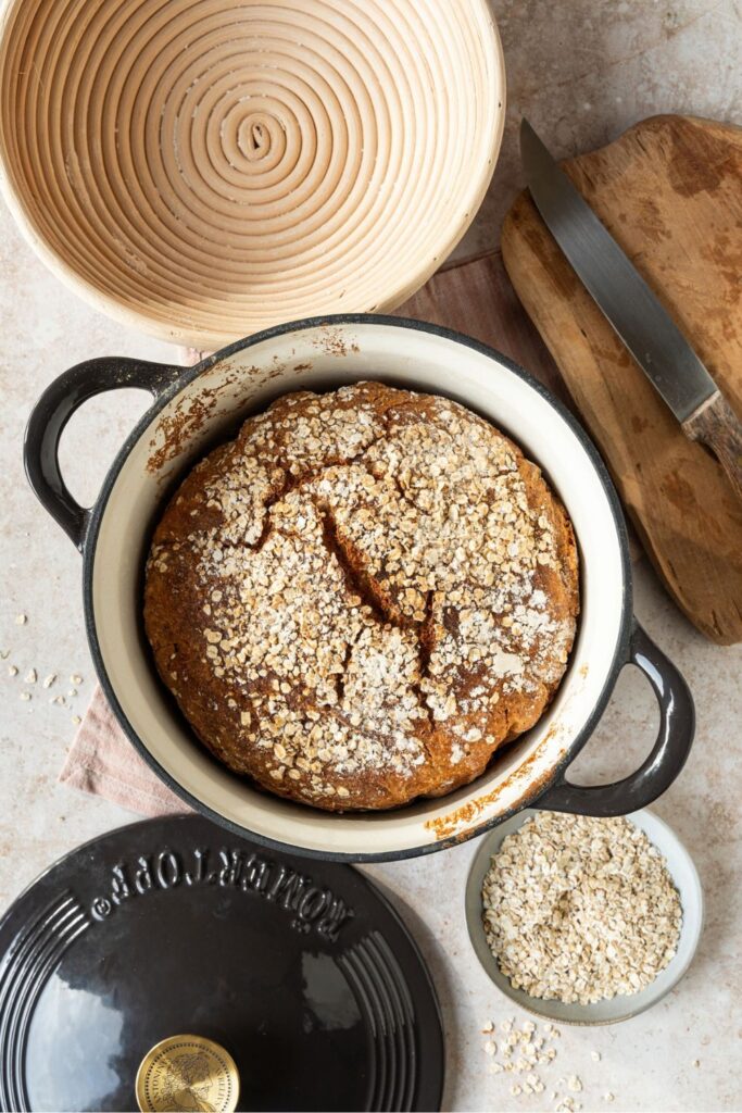 Hafer-Porridge Brot im schwarzen Gusseisentopf von oben, umgeben von Gärkörbchen und einer Schale Haferflocken