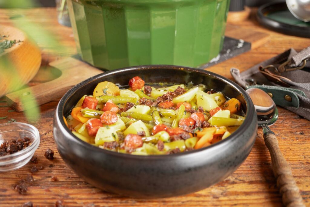 Kartoffelsuppe mit Würstchen und Pumpernickelcrunch in dunkler Schale, im Hintergrund grüner Römertopf Bräter
