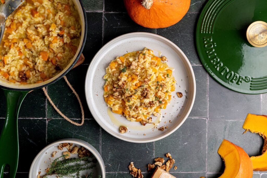 Kürbisrisotto mit Walnüssen und Thymian auf hellem Teller, im Hintergrund grüne Stielkasserolle und frischer Hokkaidokürbis