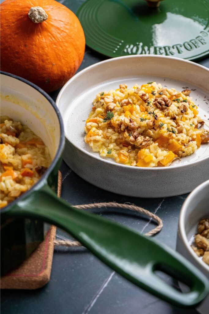 Kürbisrisotto mit Walnüssen und Thymian in heller Schale, umgeben von Hokkaidokürbis und grüner Stielkasserolle