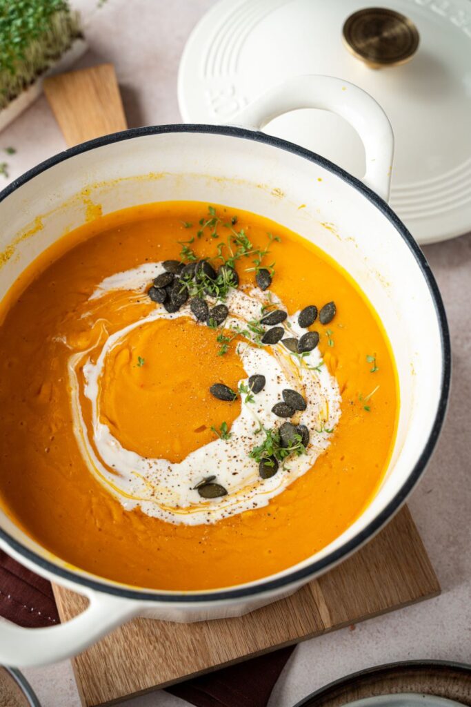 Klassische Kürbissuppe im weißen Gusseisentopf, getoppt mit Crème fraîche, Kresse und Kürbiskernen