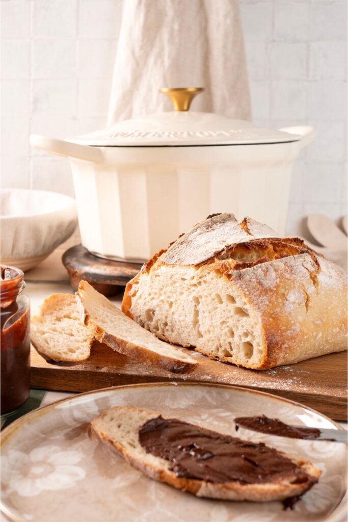 Aufgeschnittenes Lievito Madre Hausbrot vor weißem Gusseisentopf, im Vordergrund Teller mit Brotscheibe und Schok-Oh-Creme