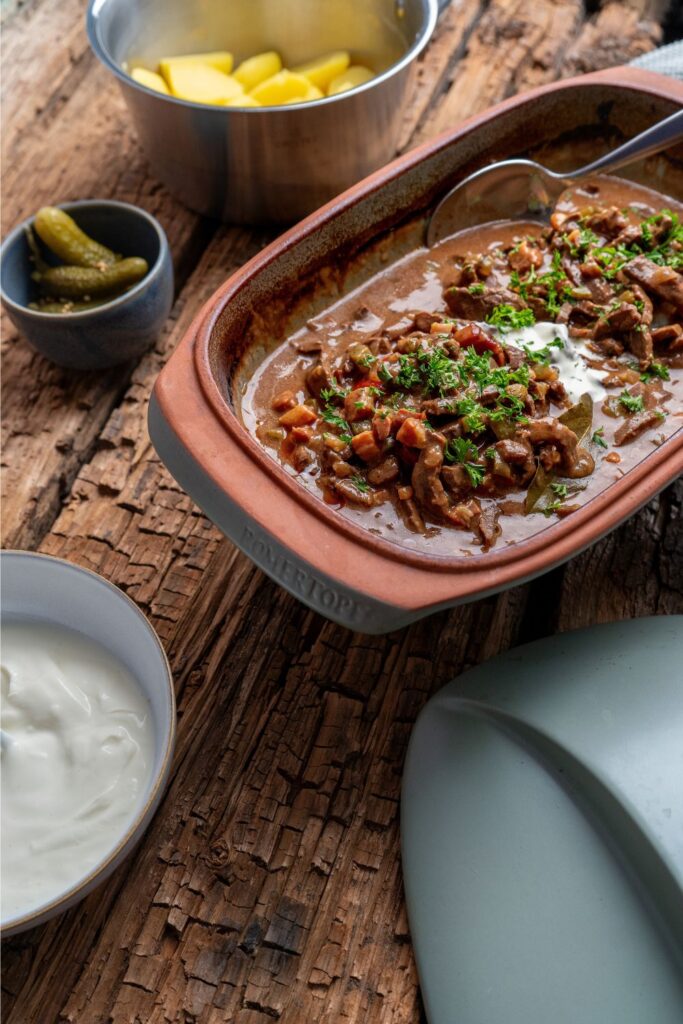 Rouladengeschnetzeltes in Römertopf-Auflaufform von oben, im Hintergrund Salzkartoffeln, Crème fraîche und Gewürzgurken