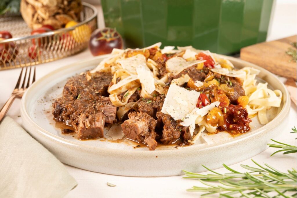 Stracotto di Manzo mit Tagliatelle, Tomatenconfit und Parmesan auf hellem Teller von Moritz und Moritz, im Hintergrund frische Tomaten