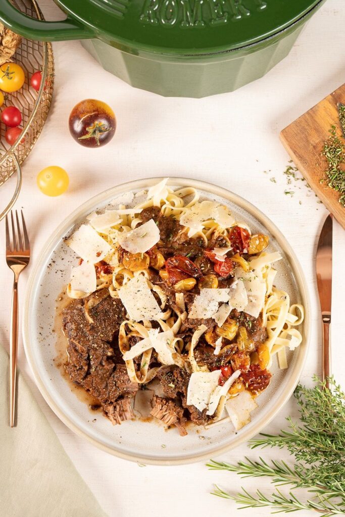 Stracotto di Manzo mit Tagliatelle und Parmesan von oben auf Teller von Moritz und Moritz, im Hintergrund grüner Gusseisentopf und Rosmarin