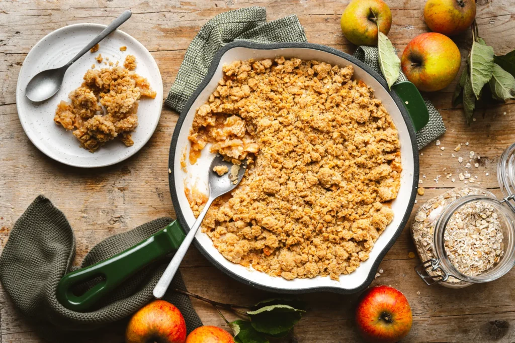 Apple Crumble mit Haferflocken-Streuseln in der grünen Gusseisenpfanne, daneben Äpfel und ein Glas Haferflocken